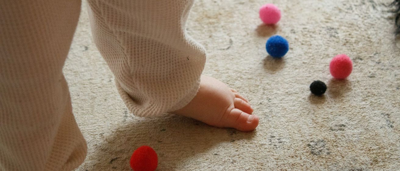 A child's foot is seen standing on a carpeted floor with various colored felt balls scattered around, including red, pink, blue, and black.
