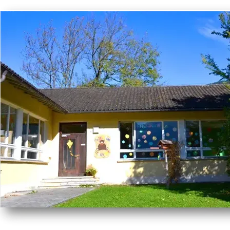 A yellow building with a brown roof, featuring a door with a decorative bear, a small garden, and large windows adorned with colorful circles.