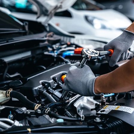 Eine Person mit Handschuhen arbeitet mit einem Schraubenschlüssel am Motor eines Autos.