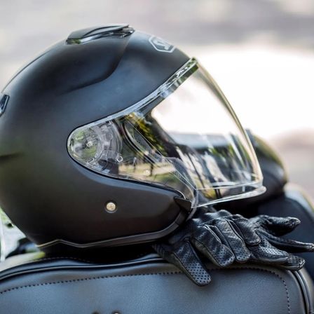 Ein schwarzer Motorradhelm mit klarem Visier sitzt auf einem schwarzen Ledersitz. Daneben liegen schwarze Motorradhandschuhe. Der Helm reflektiert einen unscharfen Hintergrund.