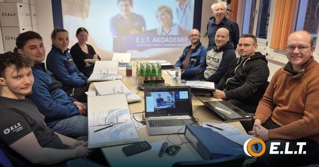 Eine Gruppe von Menschen sitzt um einen Tisch mit Laptops, Papieren und einer Projektionsleinwand. Sie scheinen in einer Besprechung zu sein.