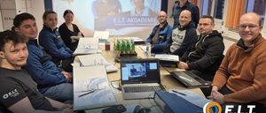 Eine Gruppe von Menschen sitzt um einen Tisch mit Laptops, Papieren und einer Projektionsleinwand. Sie scheinen in einer Besprechung zu sein.