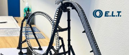 Ein schwarzes Achterbahnmodell aus Kunststoff wird auf einer weißen Oberfläche ausgestellt. Es hat mehrere Loopings und Kurven, mit verstreuten Komponenten. Das Bild hat einen Wasserzeichen von E.L.T. in der oberen rechten Ecke.