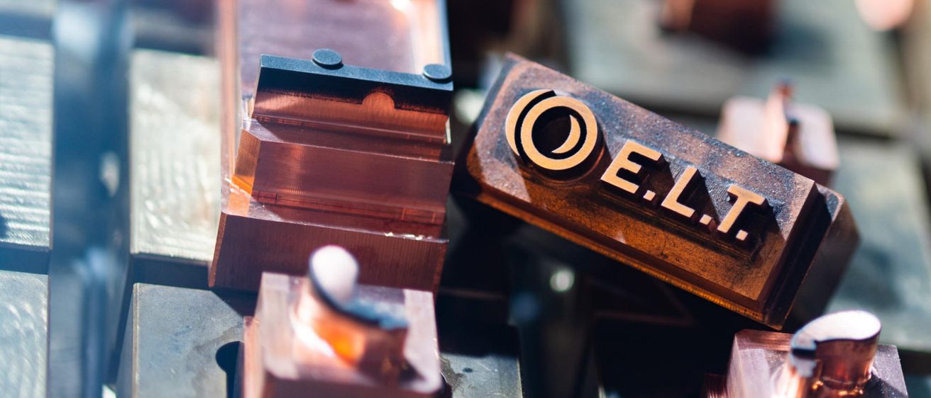 Eine Nahaufnahme von Kupferformen mit der Aufschrift O.E.L.T., Teil eines Herstellungsprozesses. Die Formen sind metallisch, kunstvoll gestaltet und in Reihen angeordnet.