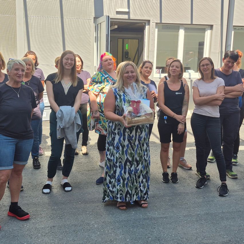 Eine Gruppe von Frauen steht vor einem Gebäude. Eine Frau hält ein verpacktes Geschenk. Einige tragen Brillen, Turnschuhe und Armbänder. Das Gebäude hat Glasfenster und eine offene Tür.