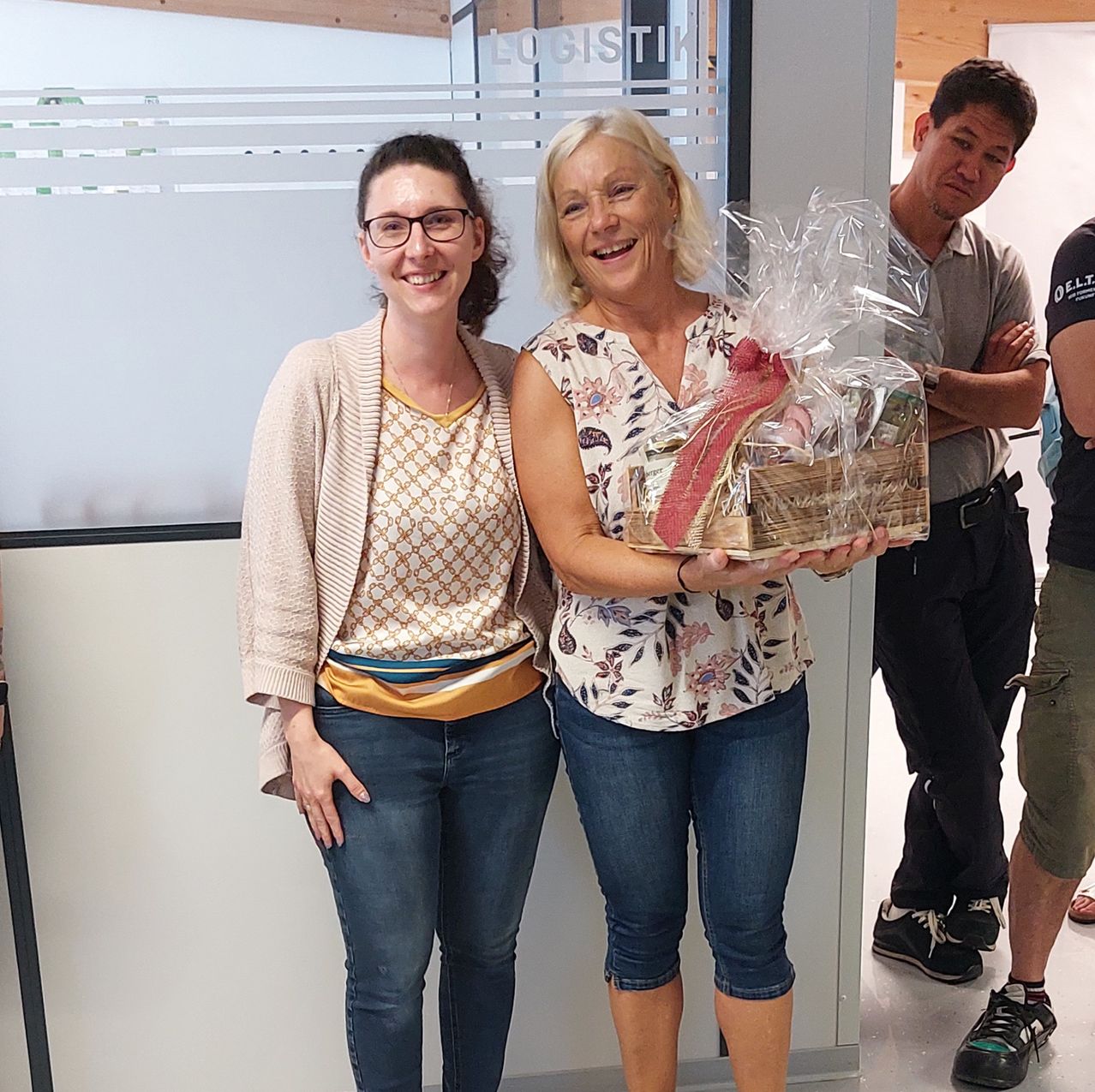 Zwei Frauen lächeln und posieren für ein Foto mit einem Geschenkkorb. Dahinter steht ein Mann mit verschränkten Armen. Das Bild wurde in einer Innenumgebung aufgenommen.