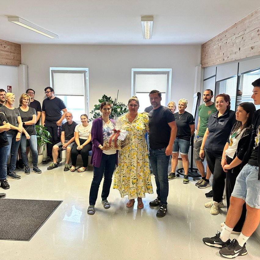 Eine Gruppe von Menschen steht in einem Raum, zwei Frauen in der Mitte halten ein Geschenk, und andere beobachten, mit Pflanzen und Fenstern im Hintergrund.