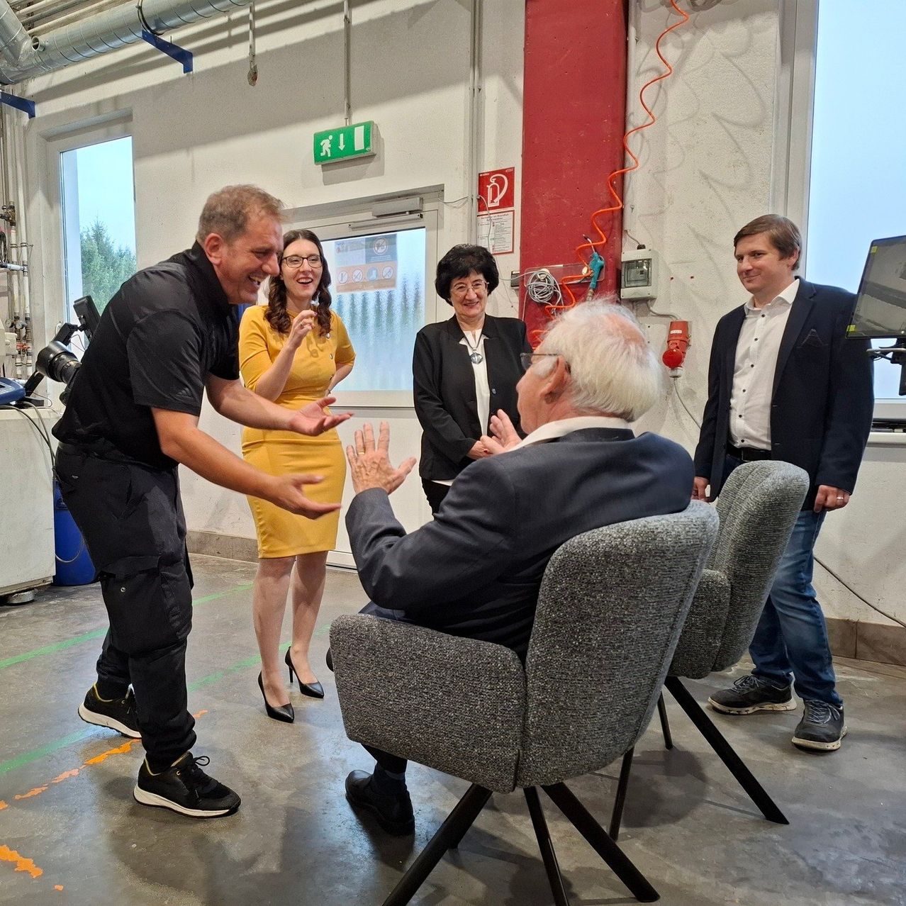 Eine Gruppe von Menschen befindet sich in einem Raum. Ein Mann sitzt auf einem Stuhl, während ein anderer Mann steht und gestikuliert. Eine Frau steht und lächelt. Ein Monitor befindet sich auf der rechten Seite.