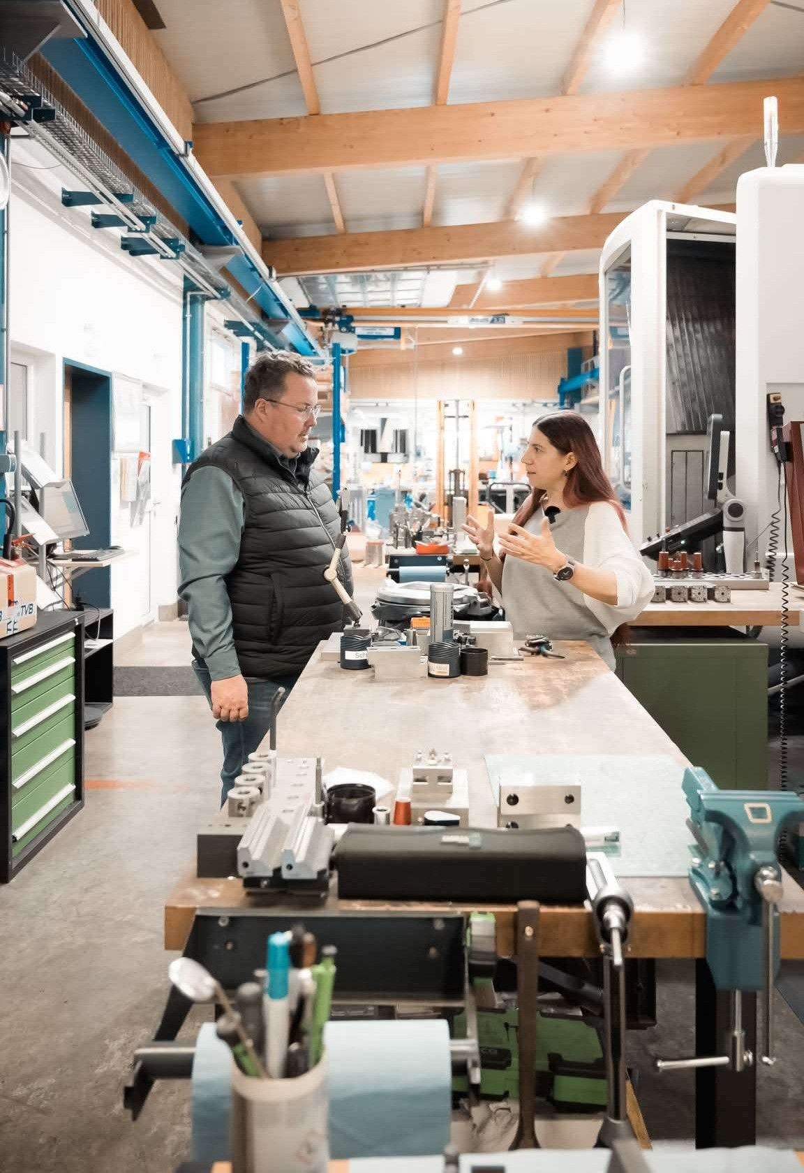 Ein Mann und eine Frau in einer Werkstatt, der Mann hält ein Werkzeug, die Frau spricht. Hinter ihnen verschiedene Maschinen und Werkzeuge.