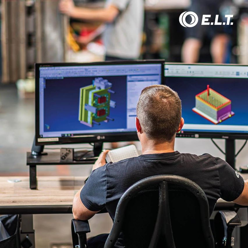 Ein Mann sitzt an einem Schreibtisch und arbeitet an einem Computer mit zwei Monitoren, die 3D-Modelle anzeigen. Er trägt ein schwarzes T-Shirt.