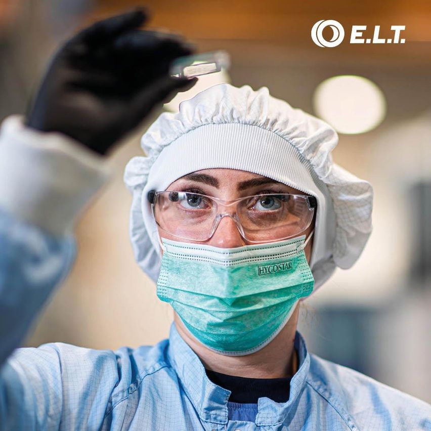 Eine Person in Laborkittel, Maske und Schutzbrille untersucht eine kleine Probe in einer Petrischale.