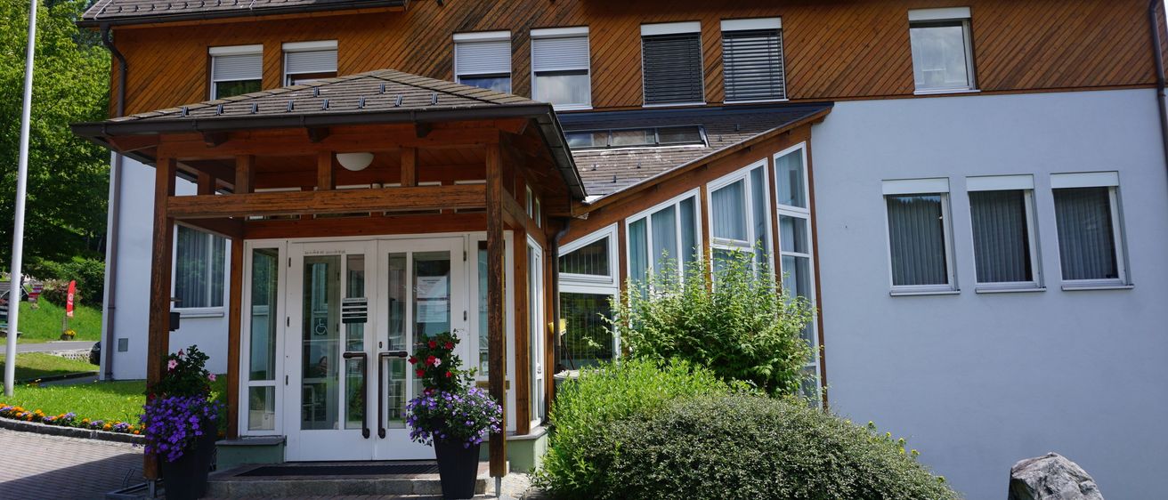 Ein braunes und weißes Gebäude mit einem gepflasterten Weg und einer überdachten Veranda. Es hat mehrere Fenster und einen Balkon mit einem kleinen Dach.