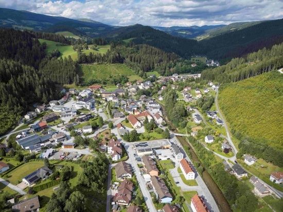 Ein Luftbild eines Dorfes mit Häusern und Gebäuden, umgeben von Bäumen und Bergen. Der Himmel ist bewölkt.