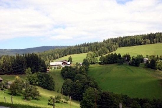 Ein weiter Blick auf eine ländliche Landschaft mit einem weißen Haus und grünen Feldern, umgeben von Bäumen und Büschen unter einem blauen, bewölkten Himmel.