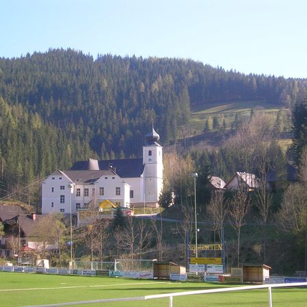 Ein weißes Gebäude mit Turm steht auf einem Hügel, umgeben von Bäumen und einem grünen Feld. Unten sind mehrere kleinere Häuser zu sehen, und Straßenlaternen säumen die Straße.