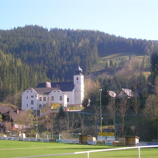 Ein weißes Gebäude mit Turm steht auf einem Hügel, umgeben von Bäumen und einem grünen Feld. Unten sind mehrere kleinere Häuser zu sehen, und Straßenlaternen säumen die Straße.