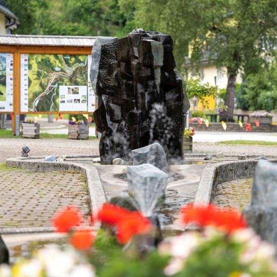 Ein felsiger Brunnen mit fließendem Wasser ist von leuchtend roten und weißen Blumen umgeben. Dahinter befindet sich eine Holzstruktur mit Karten und einem kleinen Haus. Das Gebiet ist von üppigem Grün umgeben.