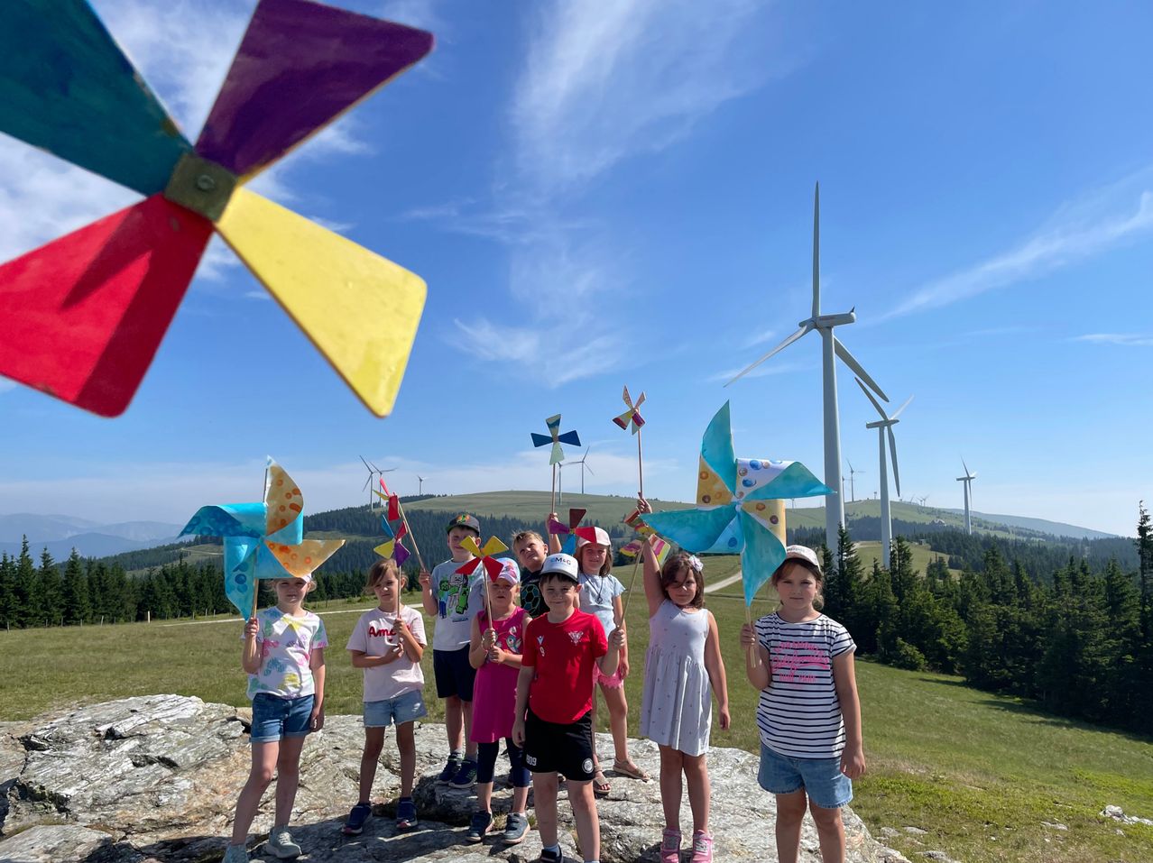 Eine Gruppe von Kindern mit bunten Windrädern steht auf einem Felsen in einem Feld mit Windrädern im Hintergrund.