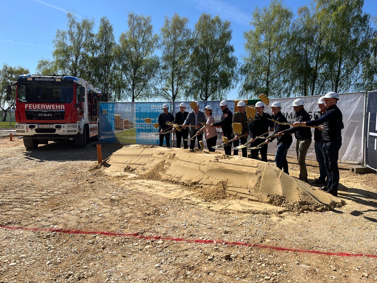 Eine Gruppe von Menschen in weißen Helmen mit Schaufeln gräbt an einer Baustelle. Ein Feuerwehrwagen und ein Banner sind im Hintergrund.