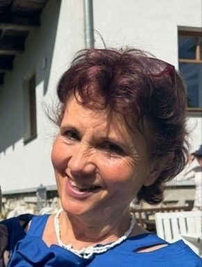Eine Frau mit kurzen, lockigen Haaren, die ein blaues Oberteil und eine weiße Perlenkette trägt, lächelt fröhlich draußen in der Nähe eines Gebäudes.