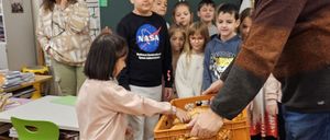 Ein Mann übergibt einem Mädchen einen Korb mit Brot in einem Klassenzimmer, während Kinder zuschauen. Eine Frau lächelt in der Nähe.
