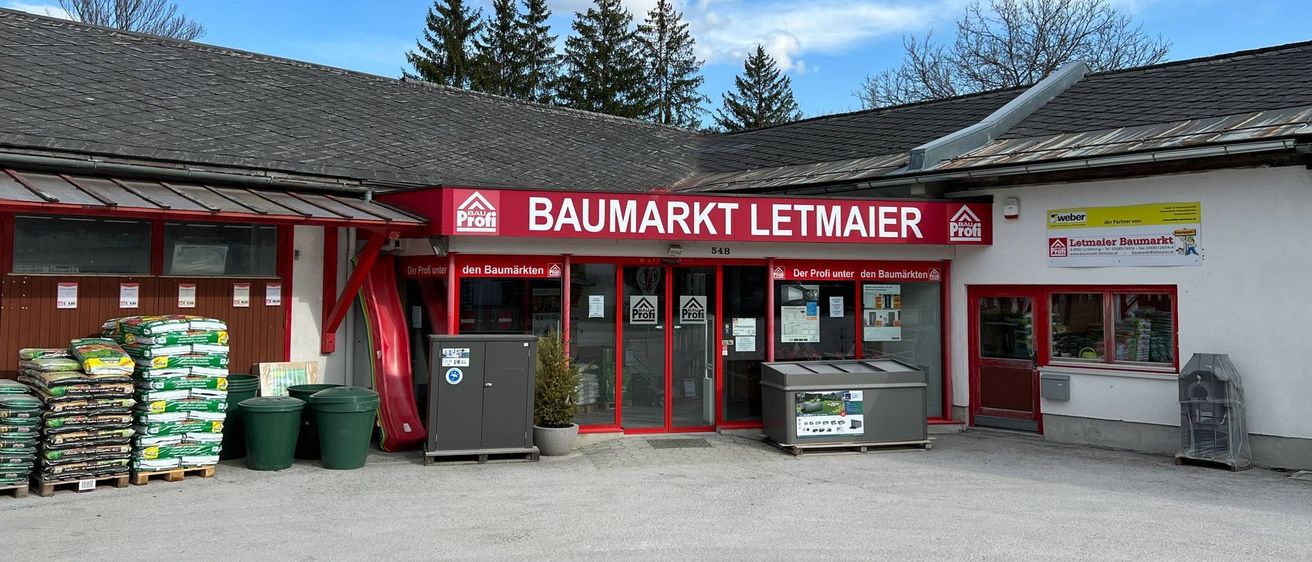 Die Vorderseite von Baumarkt Letmaier, ein Gebäude mit einem roten Schild, Glastüren und Topfpflanzen. Ein Mülleimer ist links.