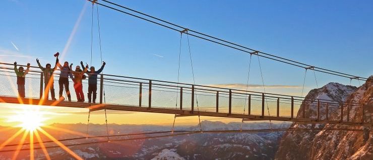 Eine Gruppe von Wanderern posiert auf einer Hängebrücke über einer verschneiten Bergkette mit einem untergehenden Sonnenuntergang im Hintergrund.