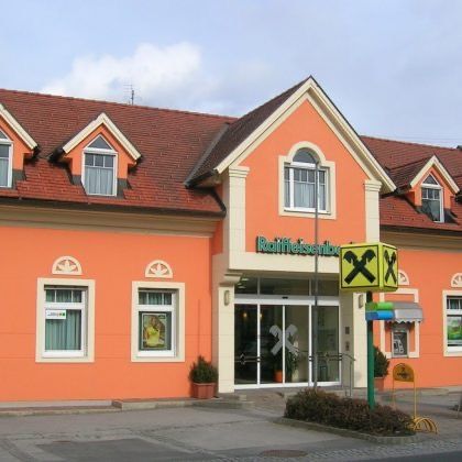 Ein orangefarbenes Gebäude mit rotem Dach und Glastüren. Es gibt Fenster, Topfpflanzen und ein gelbes Schild mit einem X darauf. Es hat das Wort 'Rathauseingang' oben.