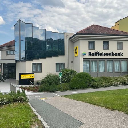 Das Äußere der Raiffeisenbank mit Glasfassaden und einem braunen Dach. Ein gelbes Schild mit schwarzem Schriftzug und Logo ist vorne angebracht.