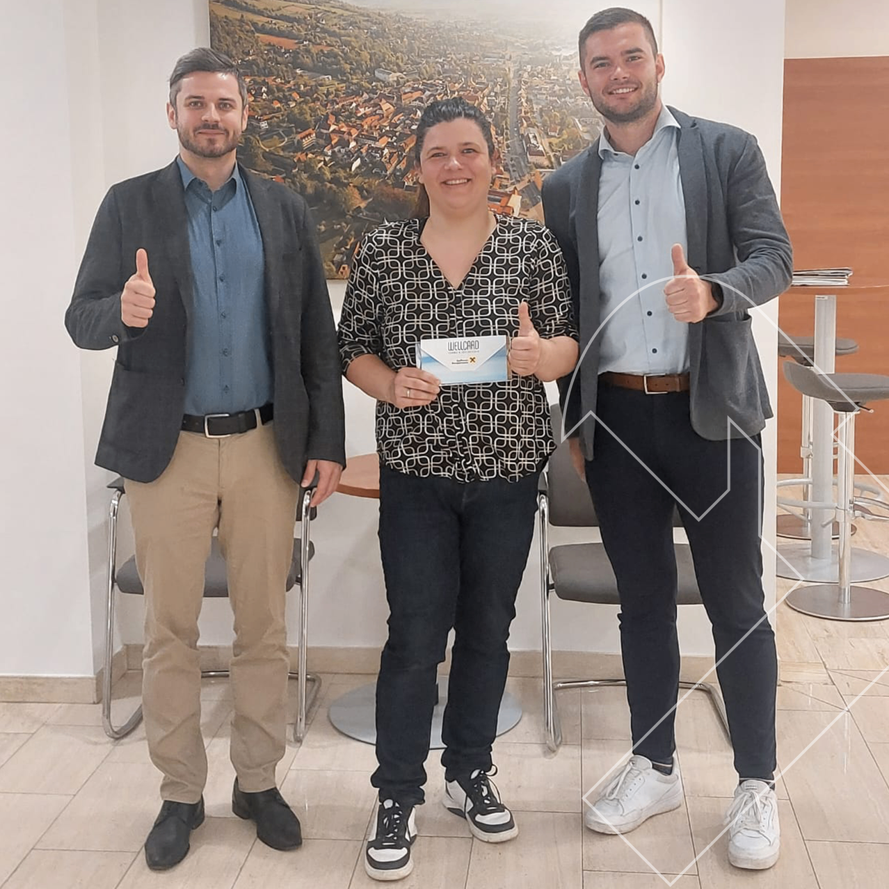 Drei Menschen stehen zusammen in einem Raum, lächeln und geben Daumen hoch. Eine Frau in der Mitte hält ein Zertifikat. Hinter ihnen ist ein Bild einer Stadt an der Wand.