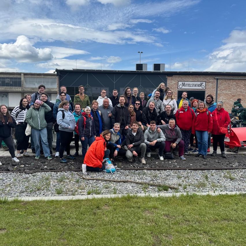 Eine Gruppe von Menschen steht auf einem Kiesweg vor einem Backsteingebäude mit einem Schild, das 'Mittelstation' besagt. Sie sind in verschiedenen Jacken gekleidet und einige tragen Brillen. Im Hintergrund befindet sich ein Gebäude mit Fenstern und einer Straßenlaterne.
