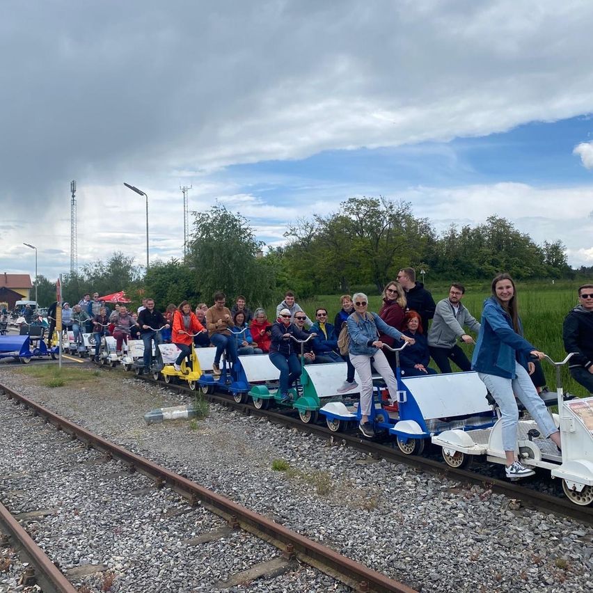 Eine Gruppe von Menschen fährt auf einem bunten Mini-Zug auf einer Eisenbahnstrecke, umgeben von Bäumen und einem bewölkten Himmel.