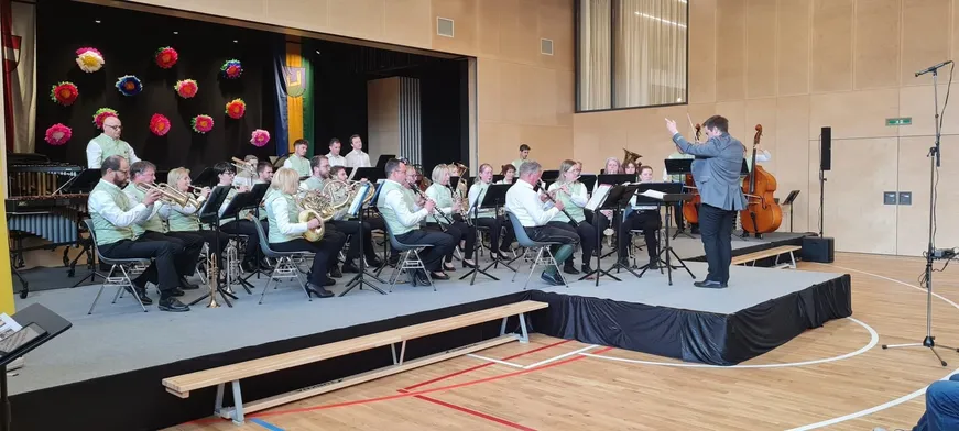 Ein Orchester probet auf der Bühne, mit Musikern, die verschiedene Instrumente wie Flöten, Trompeten und Posaunen spielen. Ein Dirigent steht vorne.