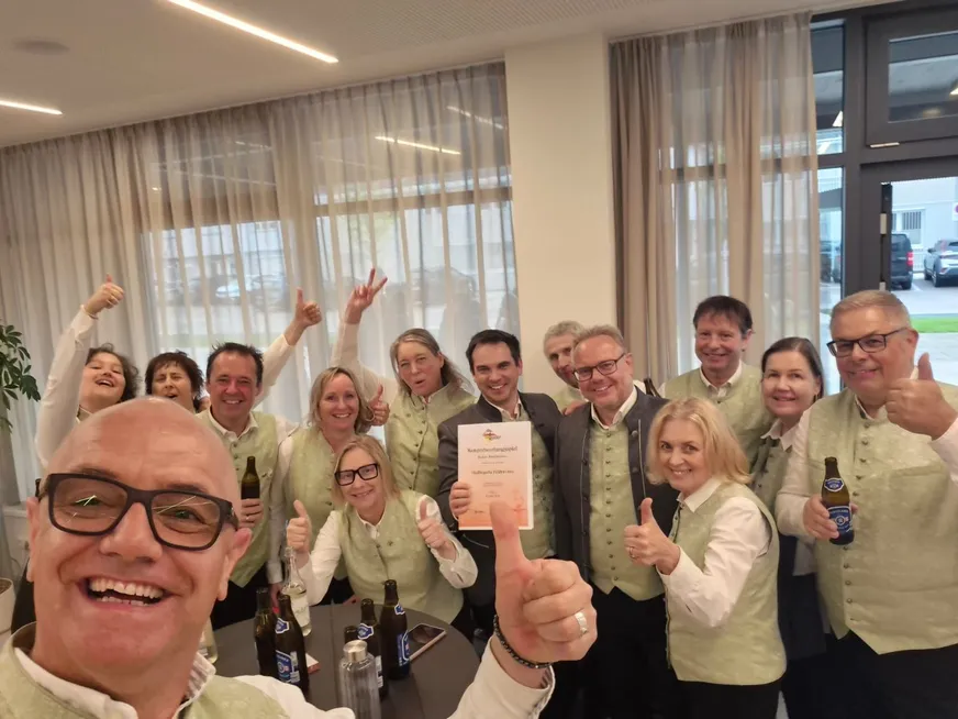 Eine Gruppe von Menschen in grünen Westen posiert für ein Foto. Ein Mann gibt ein Daumen hoch und hält eine Urkunde. Sie lächeln und scheinen etwas zu feiern. Es gibt Flaschen auf dem Tisch vor ihnen.