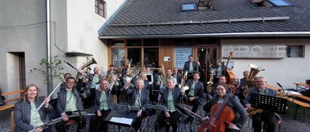Eine Gruppe von Musikern versammelt sich vor einem Gebäude und posiert für ein Foto. Sie halten verschiedene Instrumente wie Trompeten, Posaunen und Saxophone. Einige sitzen, andere stehen.