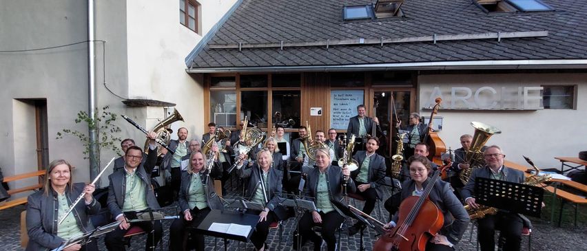 Eine Gruppe von Musikern versammelt sich vor einem Gebäude und posiert für ein Foto. Sie halten verschiedene Instrumente wie Trompeten, Posaunen und Saxophone. Einige sitzen, andere stehen.