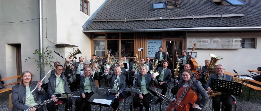 Eine Gruppe von Musikern versammelt sich vor einem Gebäude und posiert für ein Foto. Sie halten verschiedene Instrumente wie Trompeten, Posaunen und Saxophone. Einige sitzen, andere stehen.