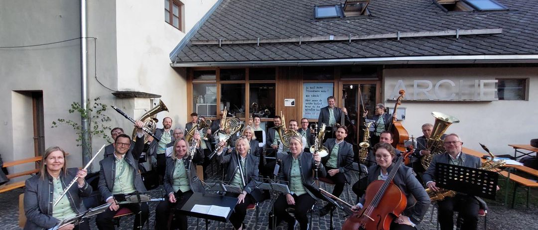 Eine Gruppe von Musikern versammelt sich vor einem Gebäude und posiert für ein Foto. Sie halten verschiedene Instrumente wie Trompeten, Posaunen und Saxophone. Einige sitzen, andere stehen.