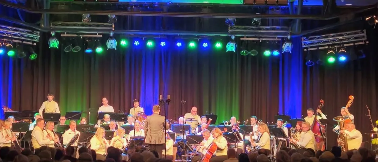Ein großes Orchester spielt auf einer Bühne mit grünen und blauen Lichtern. Ein Mann in einem Anzug steht vor dem Orchester.