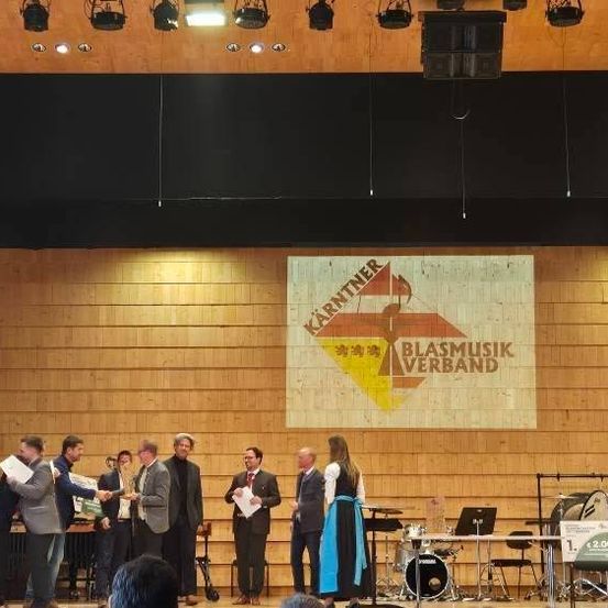 Eine Gruppe von Menschen steht auf der Bühne, einige halten Papiere, eine Person hält einen Pokal und ein Banner ist hinter ihnen. Musikinstrumente sind auf der Bühne aufgebaut.