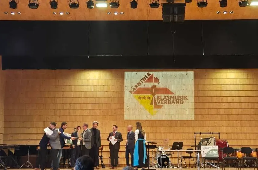 Eine Gruppe von Menschen steht auf der Bühne, einige halten Papiere, eine Person hält einen Pokal und ein Banner ist hinter ihnen. Musikinstrumente sind auf der Bühne aufgebaut.