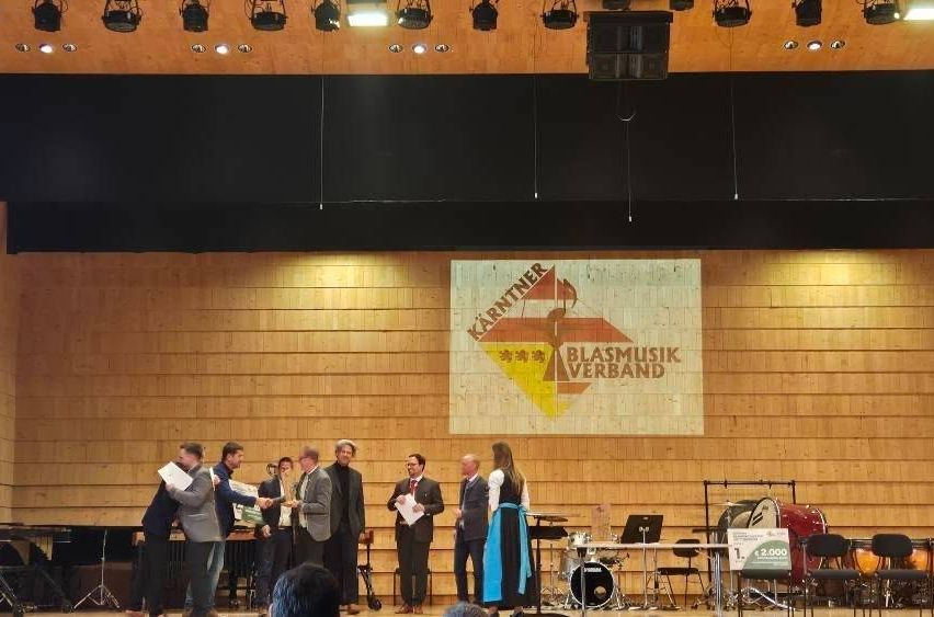 Eine Gruppe von Menschen steht auf der Bühne, einige halten Papiere, eine Person hält einen Pokal und ein Banner ist hinter ihnen. Musikinstrumente sind auf der Bühne aufgebaut.