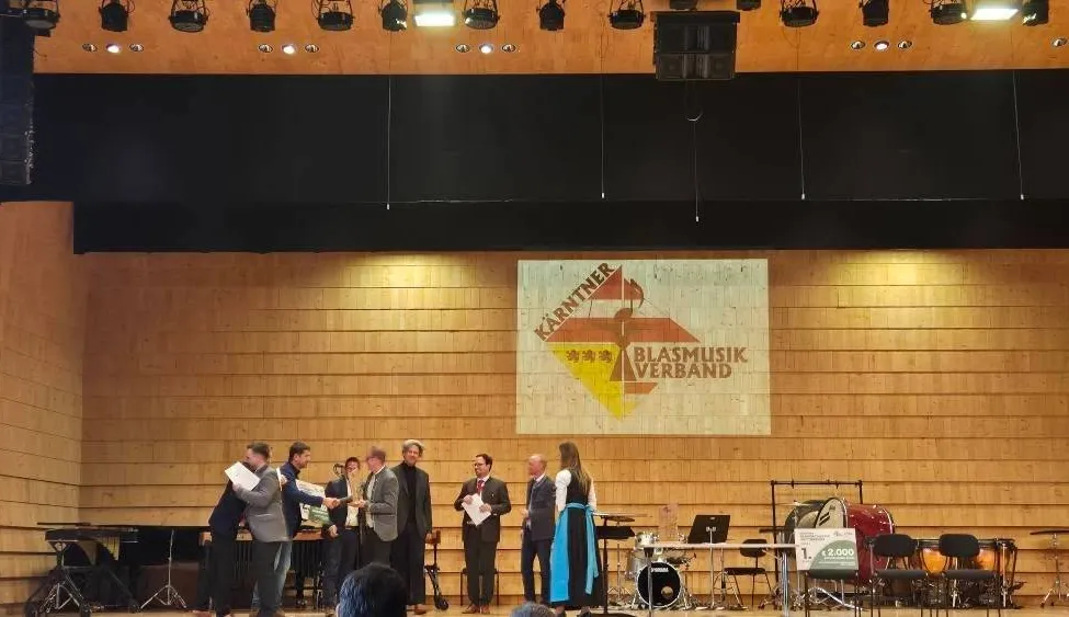 Eine Gruppe von Menschen steht auf der Bühne, einige halten Papiere, eine Person hält einen Pokal und ein Banner ist hinter ihnen. Musikinstrumente sind auf der Bühne aufgebaut.