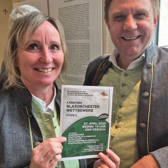 Ein Mann und eine Frau halten ein grünes Schild für einen Wettbewerb und lächeln beide in einem Raum. Dahinter ist eine weiße Wand mit Text.