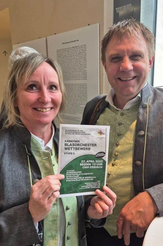 Ein Mann und eine Frau halten ein grünes Schild für einen Wettbewerb und lächeln beide in einem Raum. Dahinter ist eine weiße Wand mit Text.