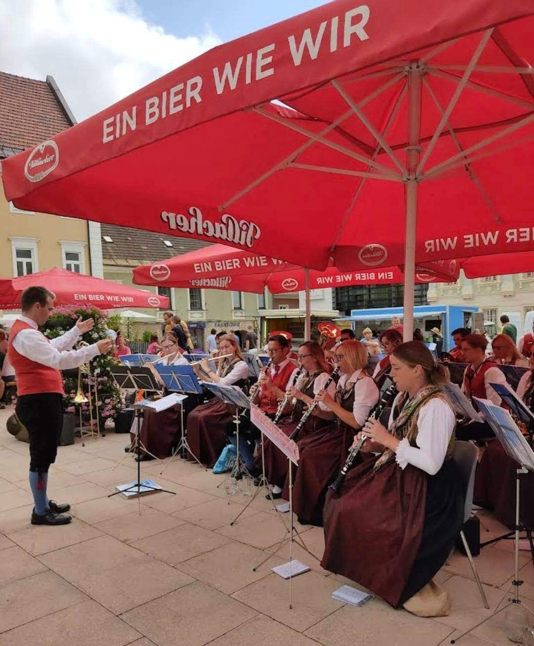 Eine Gruppe von Musikern in traditioneller Kleidung spielt auf einer Bühne mit roten Sonnenschirmen, ein Gebäude mit Balkon im Hintergrund.