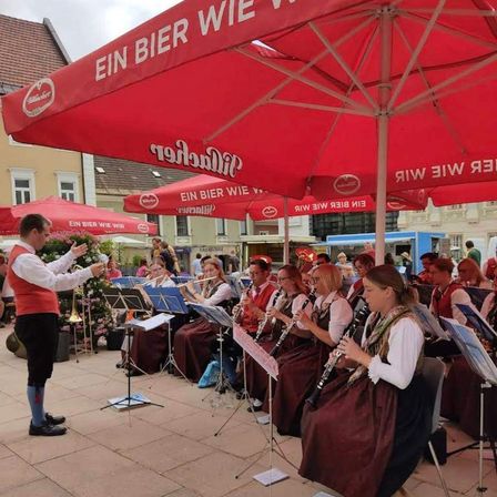 Eine Gruppe von Musikern in traditioneller Kleidung spielt auf einer Bühne mit roten Sonnenschirmen, ein Gebäude mit Balkon im Hintergrund.