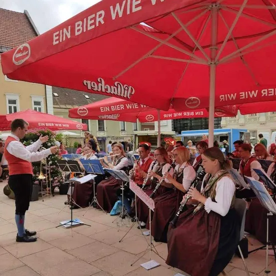 Eine Gruppe von Musikern in traditioneller Kleidung spielt auf einer Bühne mit roten Sonnenschirmen, ein Gebäude mit Balkon im Hintergrund.