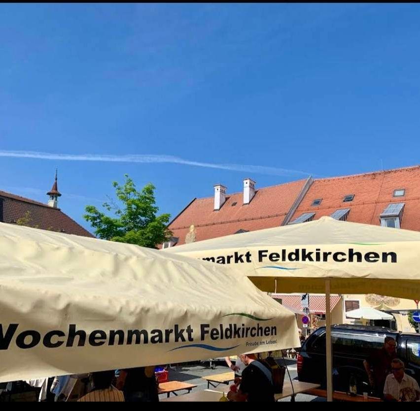 Ein Wochenmarkt unter einem weißen Baldachin, mit einem roten Gebäude im Hintergrund, einem Baum und einem klaren blauen Himmel. Menschen sitzen an Tischen unter Schirmen.