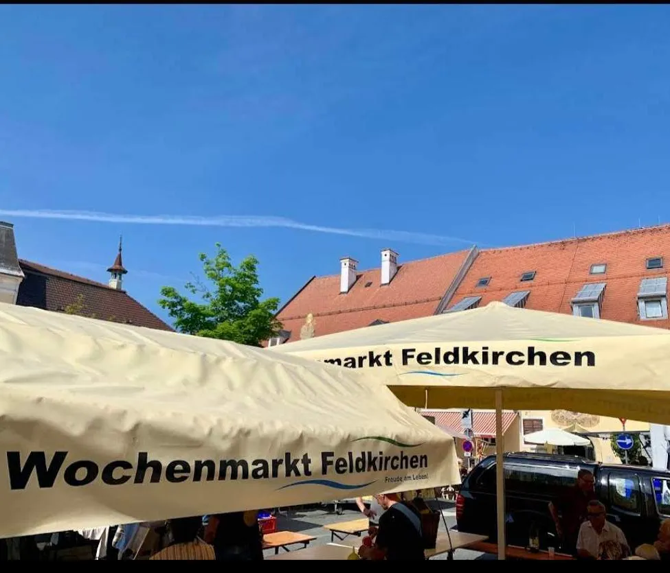 Ein Wochenmarkt unter einem weißen Baldachin, mit einem roten Gebäude im Hintergrund, einem Baum und einem klaren blauen Himmel. Menschen sitzen an Tischen unter Schirmen.